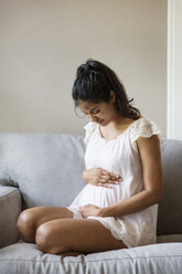 Thoughtful smiling woman touching abdomen and sitting on sofa at home - CAVF27545