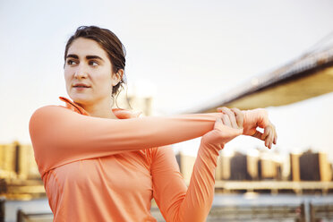 Determined female athlete stretching arm against Brooklyn Bridge - CAVF27581