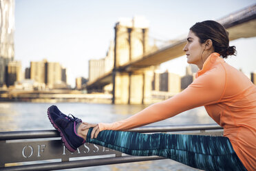 Nachdenkliche Sportlerin, die ihr Bein auf der Promenade mit der Brooklyn Bridge im Hintergrund ausstreckt - CAVF27585