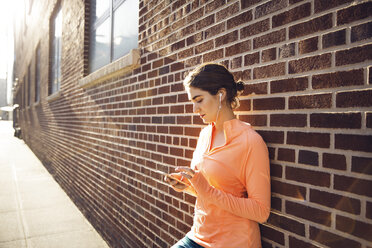 Sportliche Frau, die ihr Smartphone an einer Backsteinmauer benutzt, an einem sonnigen Tag - CAVF27588