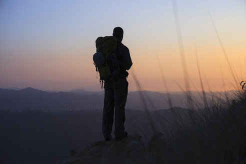 Silhouette eines Mannes, der auf einem Felsen steht und die Aussicht bei Sonnenuntergang betrachtet - CAVF27635