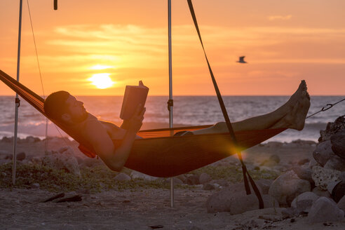 Seitenansicht eines Mannes, der in einer Hängematte am Strand bei Sonnenuntergang ein Buch liest - CAVF27670