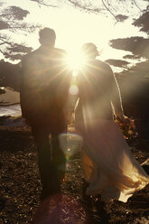 Rückansicht eines frisch vermählten Paares, das an einem sonnigen Tag auf einem Feld spazieren geht - CAVF27708