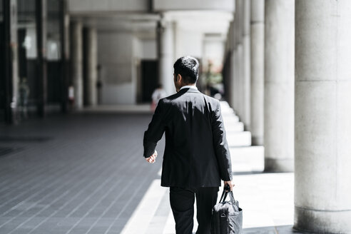 Rückansicht eines Geschäftsmannes, der eine Aktentasche trägt, während er auf einer Kolonnade geht - CAVF27745