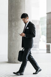Geschäftsmann in voller Länge, der ein Smartphone benutzt, während er auf der Kolonnade spazieren geht - CAVF27746