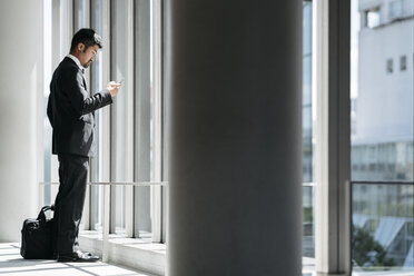 Seitenansicht eines Geschäftsmannes, der ein Smartphone benutzt, während er am Fenster im Büro steht - CAVF27747