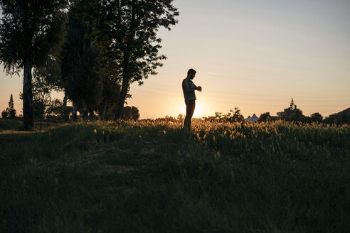 Mann, der einen Tracker untersucht, während er auf einem grasbewachsenen Feld gegen den klaren Himmel bei Sonnenuntergang steht - CAVF27761