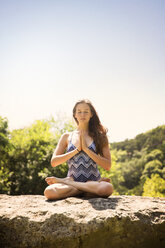 Teenager meditating on top of rock against clear sky at forest - CAVF27885