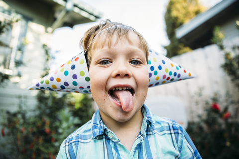 Porträt eines glücklichen Jungen mit Partyhut, der seine Zunge herausstreckt, während er im Hinterhof steht, lizenzfreies Stockfoto