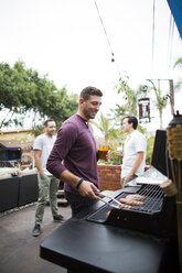 Man preparing food on barbecue grill while standing with friends in background - CAVF28006