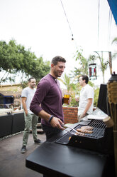 Mann bereitet Essen auf dem Grill zu, während er mit Freunden im Hintergrund steht - CAVF28006