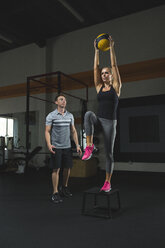 Trainerin betrachtet Frau, die einen Fitnessball hält und auf einem Hocker im Fitnessstudio steht - CAVF28038