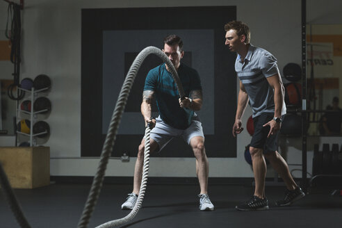 Trainer schaut auf einen männlichen Kunden, der mit Seilen im Fitnessstudio trainiert - CAVF28039