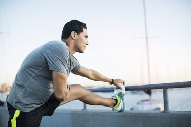 Thoughtful male athlete stretching leg on pier by harbor - CAVF28211