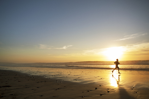 Seitenansicht eines männlichen Sportlers, der bei Sonnenuntergang am Strand läuft, lizenzfreies Stockfoto