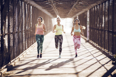 Sportlerinnen in voller Länge beim Joggen auf einer überdachten Brücke an einem sonnigen Tag - CAVF28303