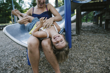 Mother playing with daughter while sitting on slide at park - CAVF28344