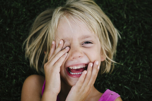 Overhead-Porträt eines glücklichen Mädchens, das sich die Augen reibt, während es im Gras liegt - CAVF28363