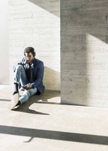 Businessman sitting on the floor in sunshine wearing headphones looking at cell phone stock photo