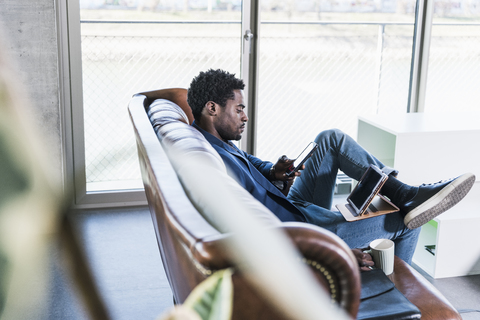 Geschäftsmann sitzt auf der Couch und benutzt Handy und Tablet, lizenzfreies Stockfoto