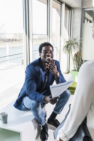 Smiling businessman with papers sitting at the window meeting colleague stock photo