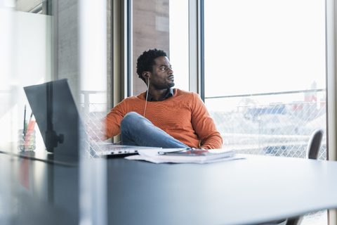 Casual businessman with earphones sitting at desk looking out of window stock photo