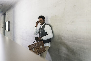 Businessman standing at concrete wall with earphones, cell phone and briefcase - UUF13122