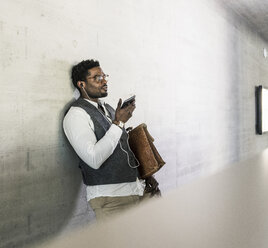 Businessman standing at concrete wall with earphones, cell phone and briefcase - UUF13123