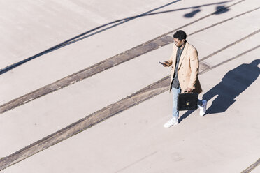 Businessman walking outdoors with briefcase, cell phone and earphones - UUF13137