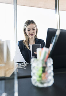 Lächelnde junge Geschäftsfrau mit Handy am Schreibtisch im Büro - UUF13156