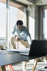 Lässiger Geschäftsmann mit Laptop, der an einem Tisch im Büro an einem Plan arbeitet - UUF13162