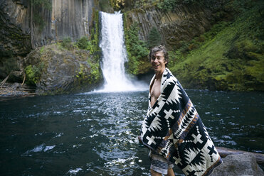 Porträt eines glücklichen Mannes mit Schal an einem Wasserfall im Wald - CAVF28430