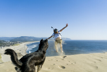 Verspielter Mann springt, während Hund am Strand gegen den klaren blauen Himmel steht - CAVF28434