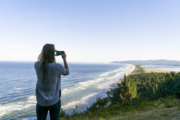 Rückansicht einer Frau, die das Meer fotografiert, während sie vor einem klaren Himmel steht - CAVF28441