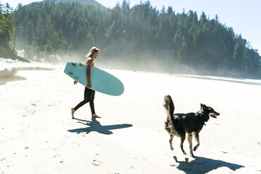 Frau hält Surfbrett mit Hund, der auf Sand am Strand spazieren geht - CAVF28444