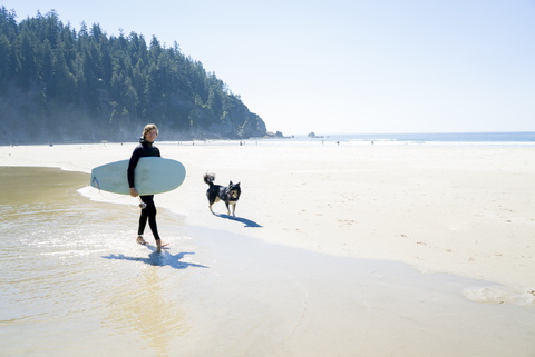 Porträt eines glücklichen Mannes, der ein Surfbrett hält und mit seinem Hund am Strand spazieren geht, lizenzfreies Stockfoto