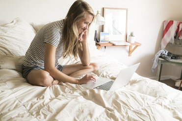 Lächelnde Frau, die einen Laptop benutzt, während sie auf dem Bett sitzt - CAVF28456