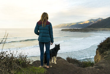 Rear view of woman with dog standing on hill against sea - CAVF28470