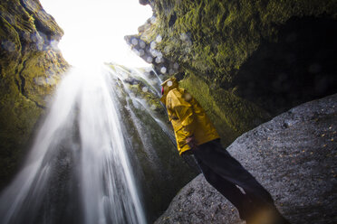 Low angle view of man wearing raincoat while looking at waterfall - CAVF28484