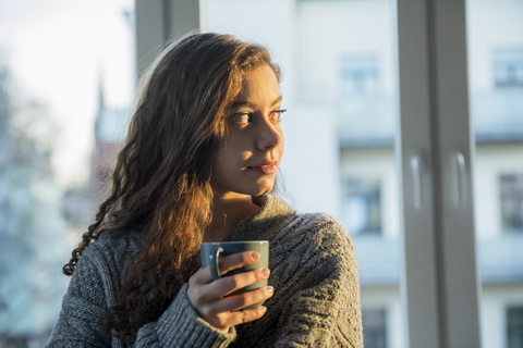 Portrait of teenage girl with coffee mug looking out of window in the evening stock photo