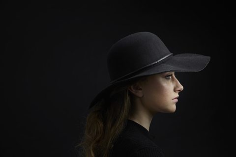 Profil einer traurigen jungen Frau mit schwarzem Hut vor schwarzem Hintergrund, lizenzfreies Stockfoto