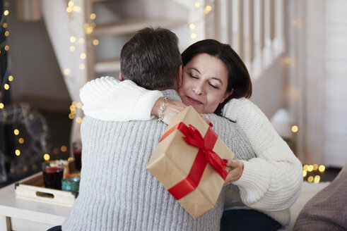 Glückliches reifes Paar mit Geschenkbox umarmt im Wohnzimmer - ABIF00197