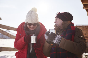 Happy mature couple with hot drinks outdoors at mountain hut in winter - ABIF00202