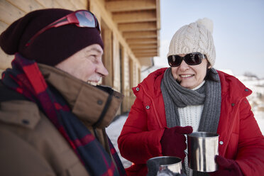Happy mature couple with hot drinks outdoors at mountain hut in winter - ABIF00204