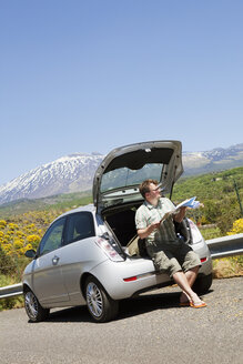 Mid adult man sitting in car trunk holding map - FOLF00736