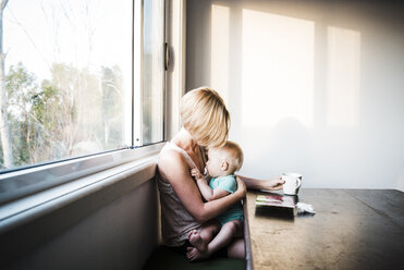 Tochter saugt am Schnuller, während sie auf dem Schoß der Mutter am Fenster sitzt - CAVF28540