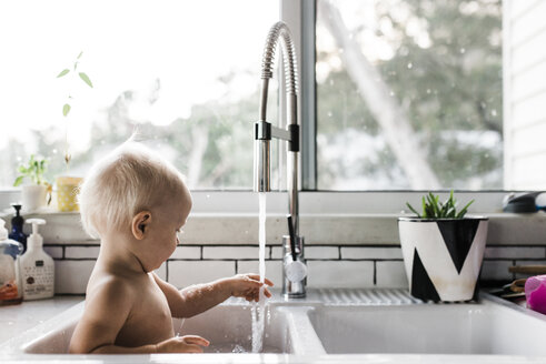 Seitenansicht eines Kleinkindes ohne Hemd, das mit Wasser spielt, während es zu Hause in der Spüle sitzt - CAVF28546