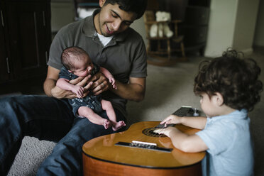 Baby boy playing guitar while sitting by father and newborn brother at home - CAVF28564