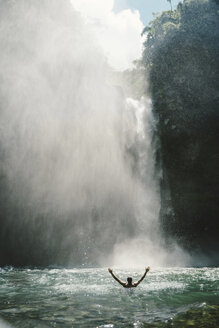 Rückansicht eines Mannes mit ausgestreckten Armen im Fluss bei einem majestätischen Wasserfall - CAVF28613