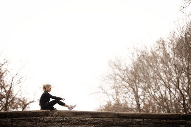 Woman sitting on retaining wall against clear sky - CAVF28660